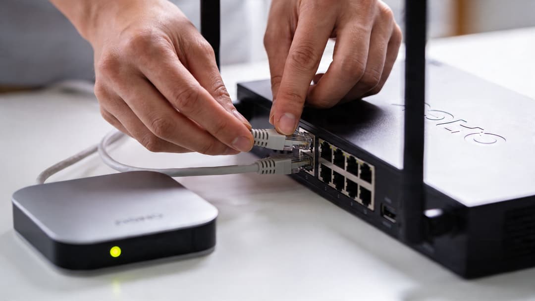 Uma pessoa conectando um cabo Ethernet em um switch de rede preto conectado a um pequeno roteador.